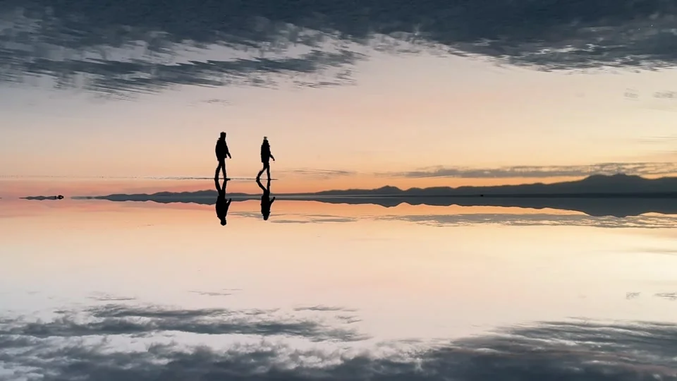 Two people walking on the mirror-like Uyuni Salt Flats at sunset